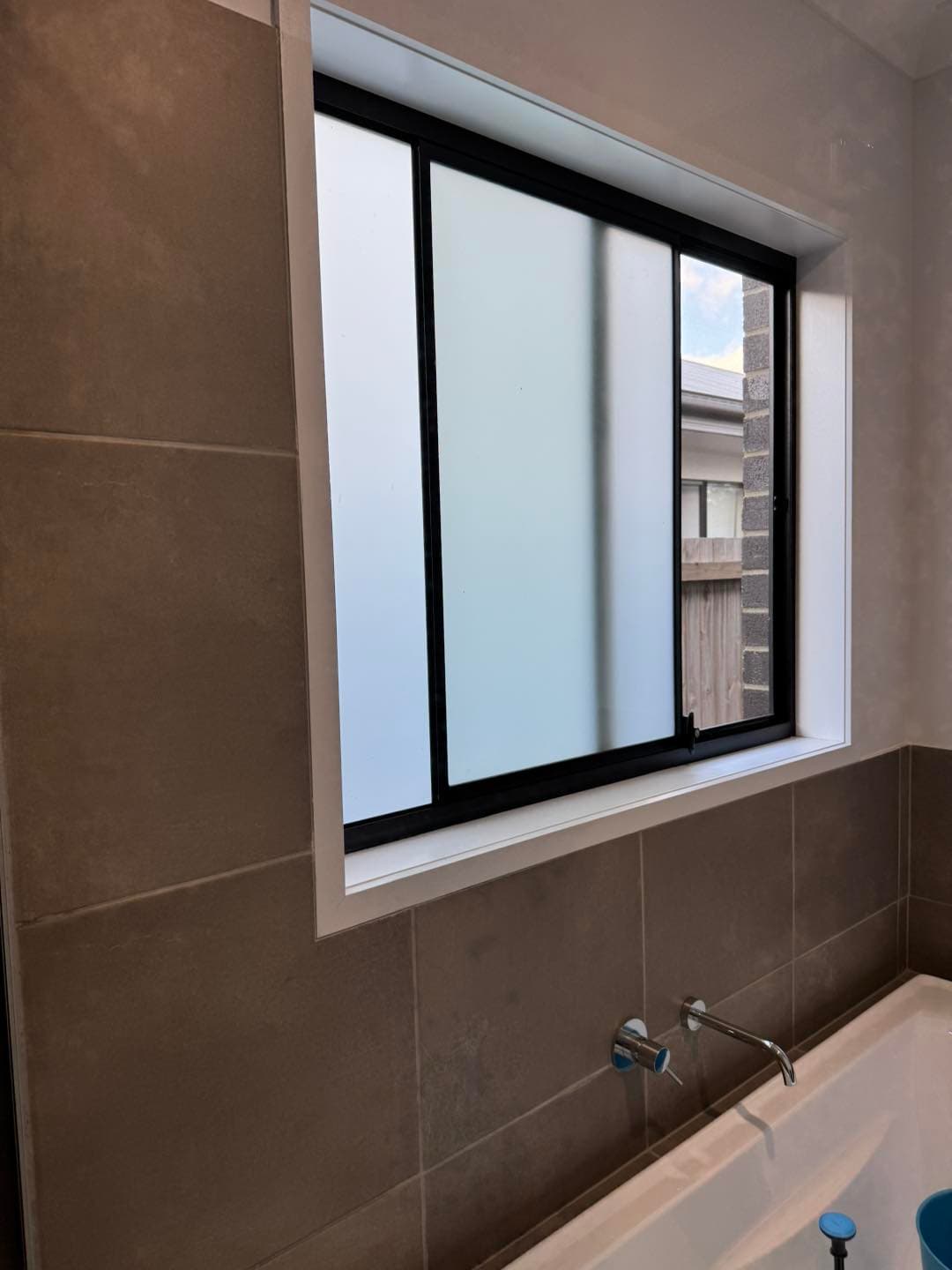 Frosted glass window with black frame set in a tiled wall above a white bathtub.