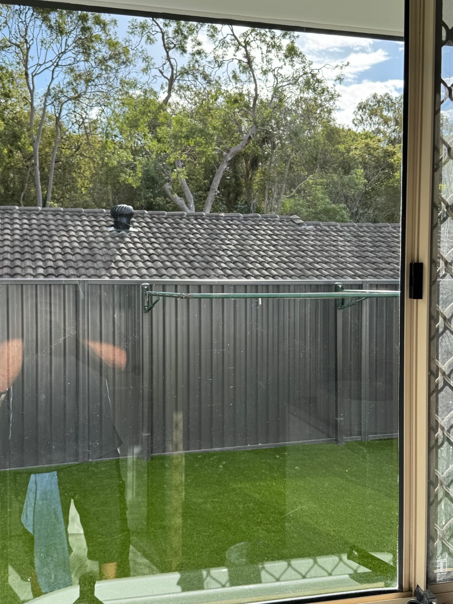 View through a window of a green lawn, grey fence, and neighboring tiled roof.